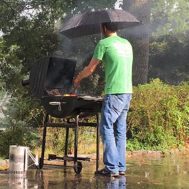 James McMenamin cooking in a green t-shirt and blue jeans wearing a black umbrella.
